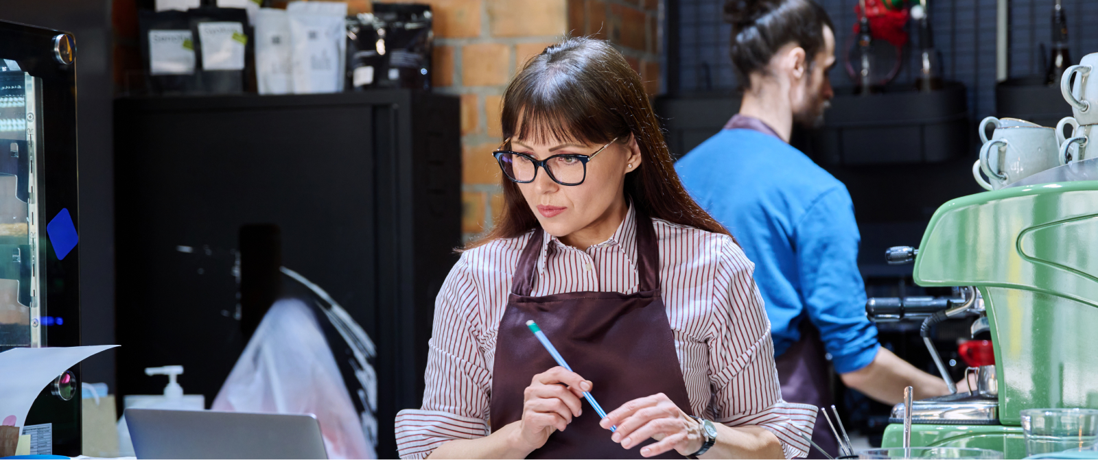 A woman in an apron reviews data on a laptop, exemplifying leadership and systems thinking in a cafe setting.