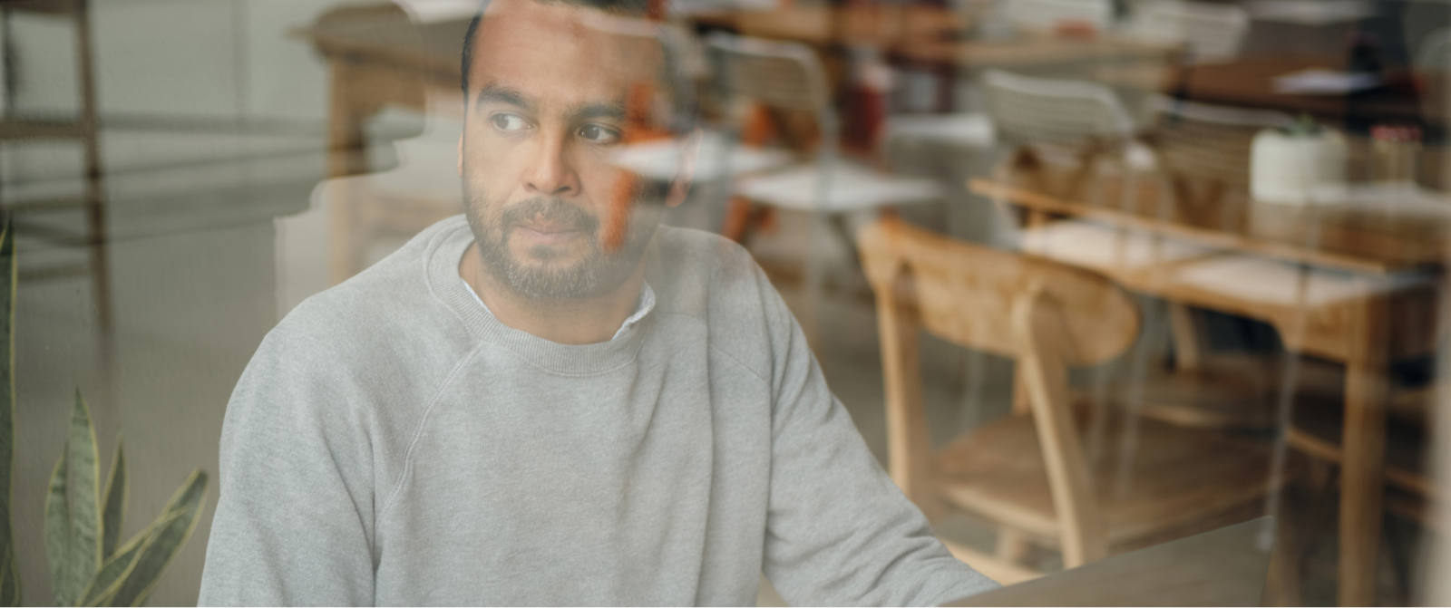 A bearded man in a gray sweatshirt sits indoors, reflecting on leadership and scaling amid quiet, empty tables outside.