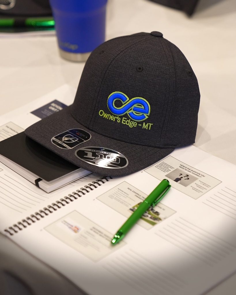 Owners Edge MT baseball cap on a professional workbook with a pen, representing intentional leadership and growth.