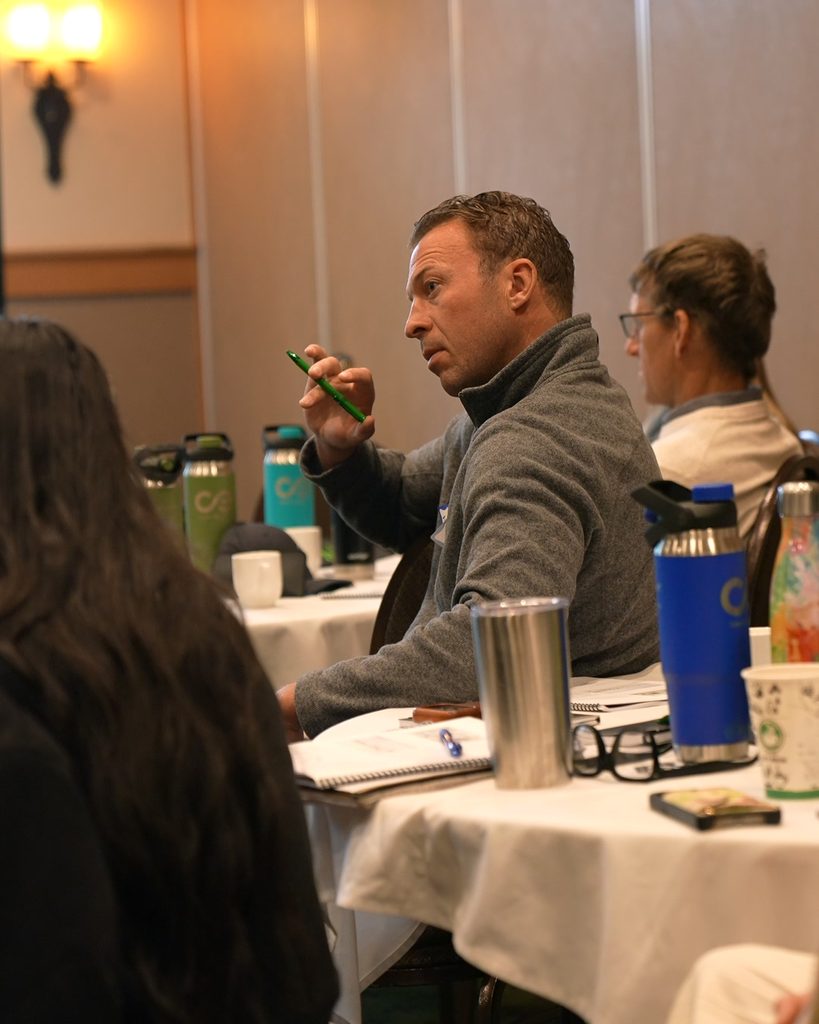A leader in a grey sweater gestures with a pen, fostering team alignment and systems thinking during a strategic meeting.