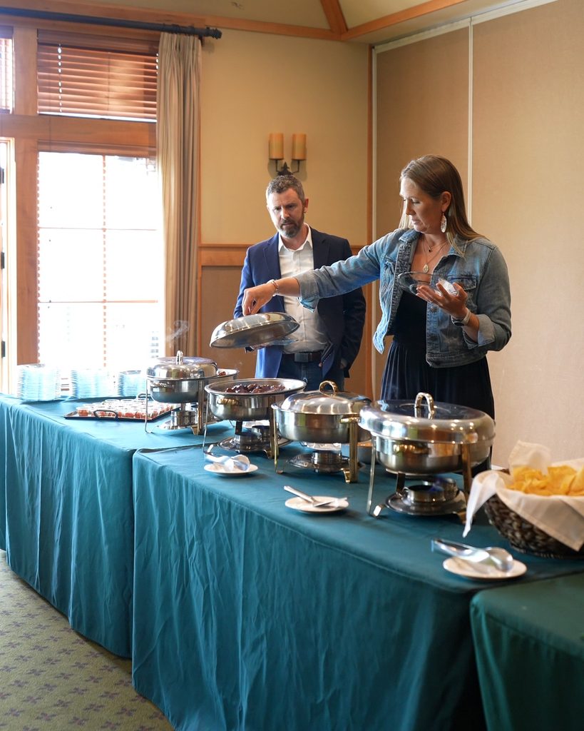 A woman in a denim jacket opens a buffet dish as a man in a blazer observes, symbolizing collaborative leadership.