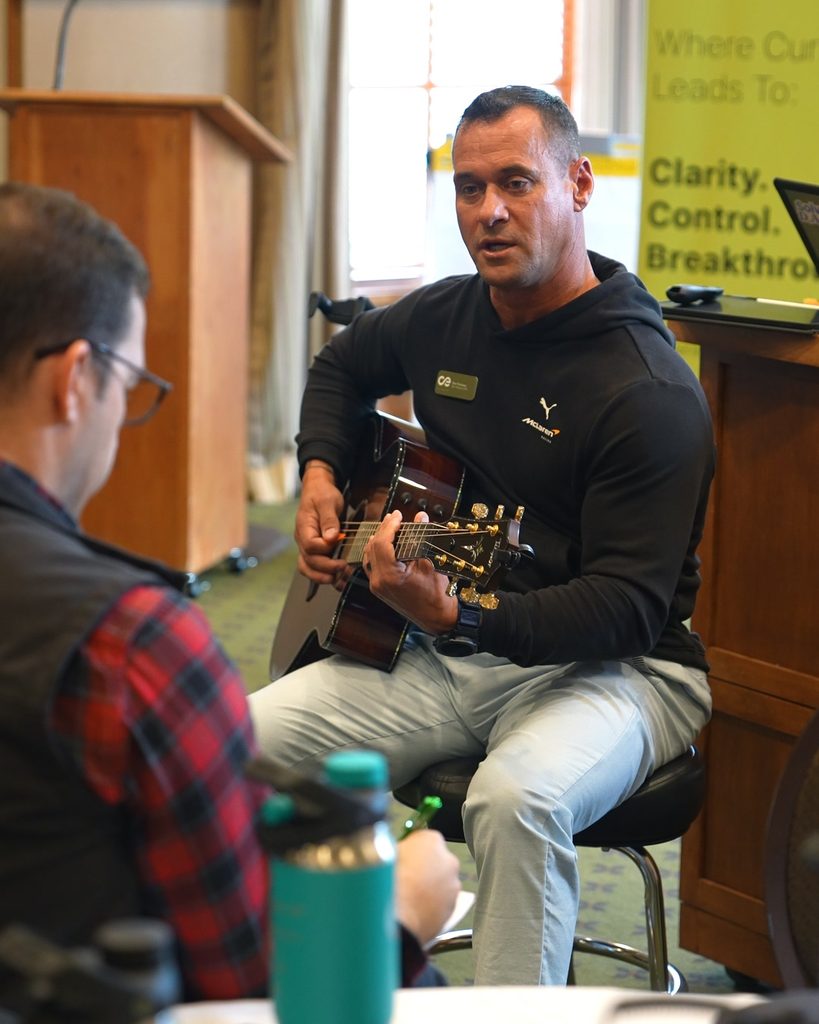 A professional in a hoodie plays guitar while discussing organizational alignment and sustainable growth with a colleague.