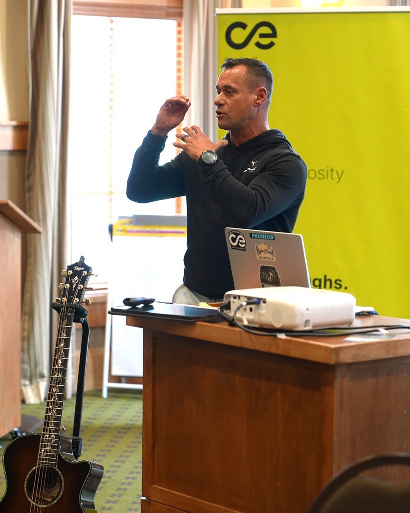 A speaker in a black hoodie shares insights on scaling and systems thinking at a podium with a laptop and guitar nearby.