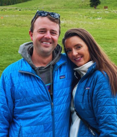 A smiling man and woman in blue jackets stand together outdoors, reflecting leadership alignment amid a scenic landscape.
