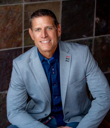 A confident leader in a gray blazer and blue shirt smiles, reflecting alignment and sustainable business growth.